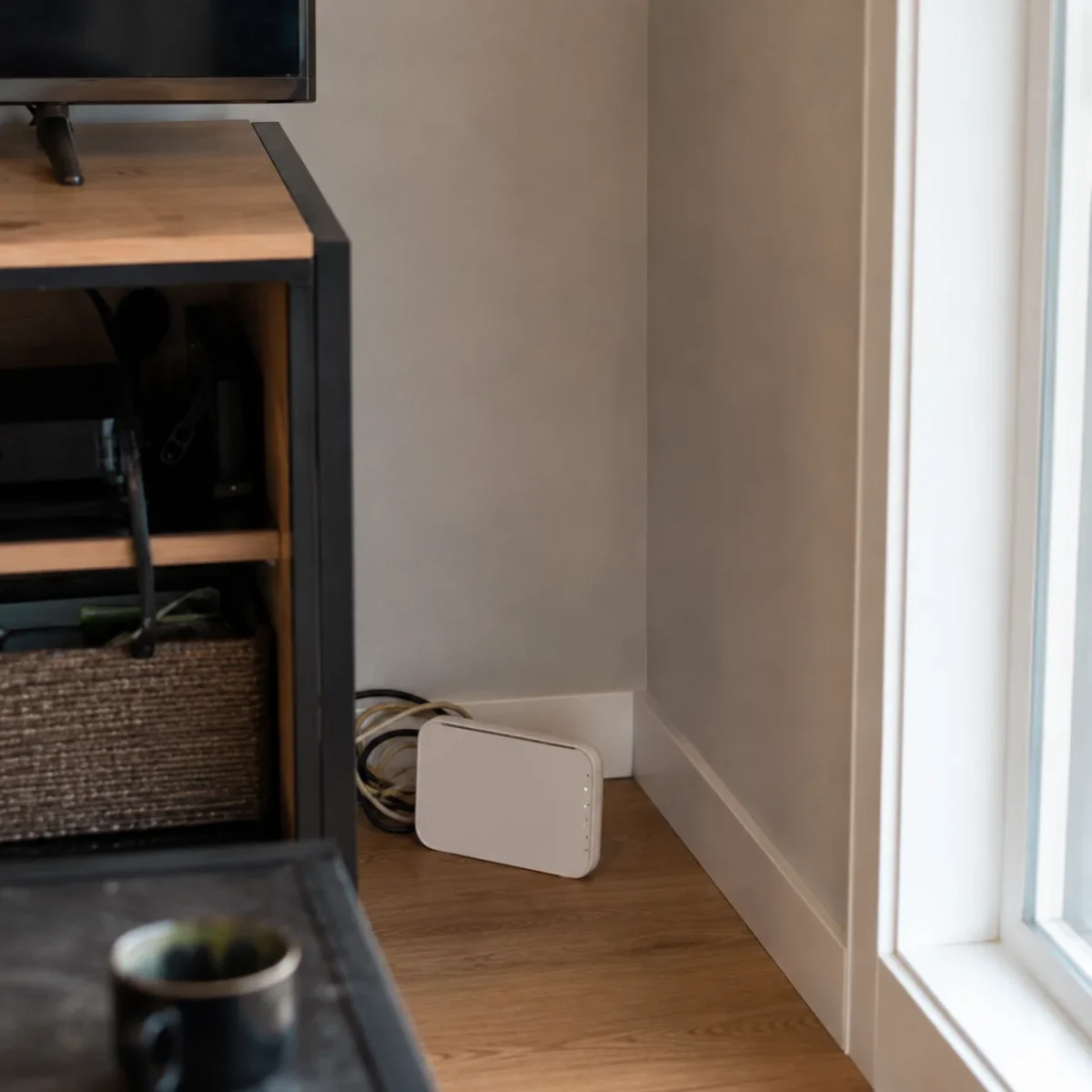 Router Wi‑Fi ustawiony w kącie pokoju, nisko przy podłodze obok szafki RTV i okna.