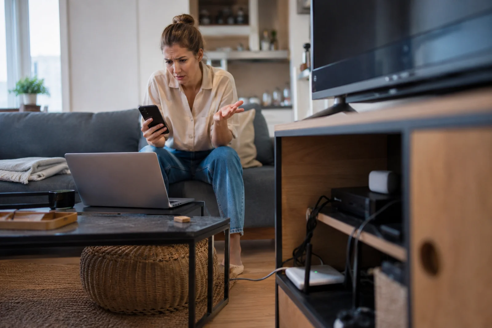 Kobieta siedzi w salonie z telefonem i laptopem, a router Wi‑Fi stoi nisko przy szafce RTV, co może powodować słaby zasięg internetu.